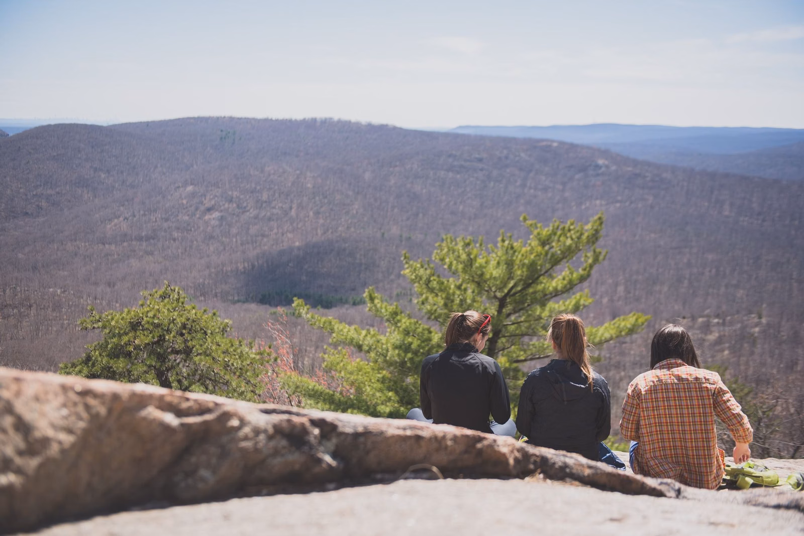 Three hikers seen from behind resting on a rock ledge above wooded Hudson Highlands ridges