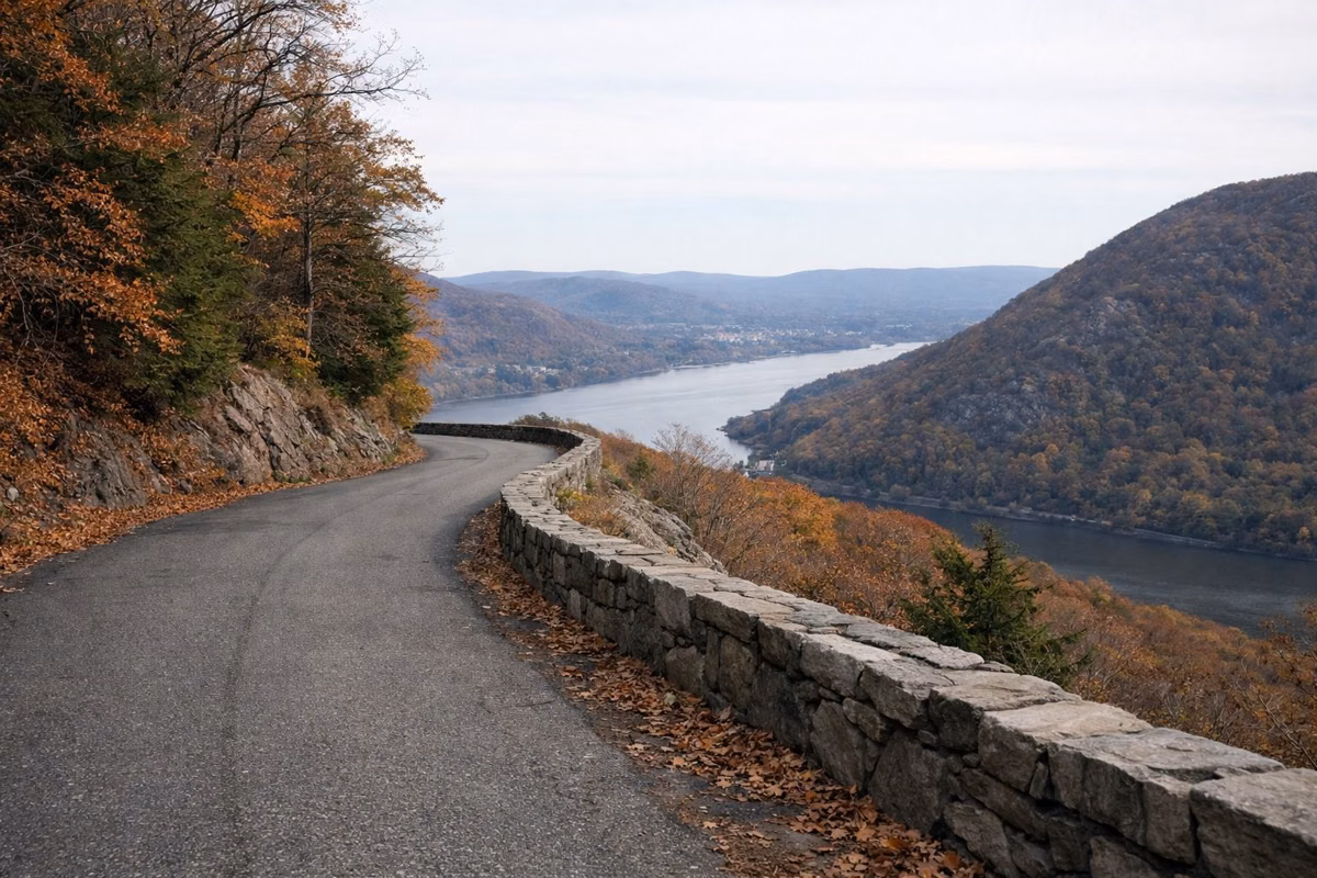 A narrow paved mountain road with a low stone wall winding up a forested summit, wide valley view beyond, no cars