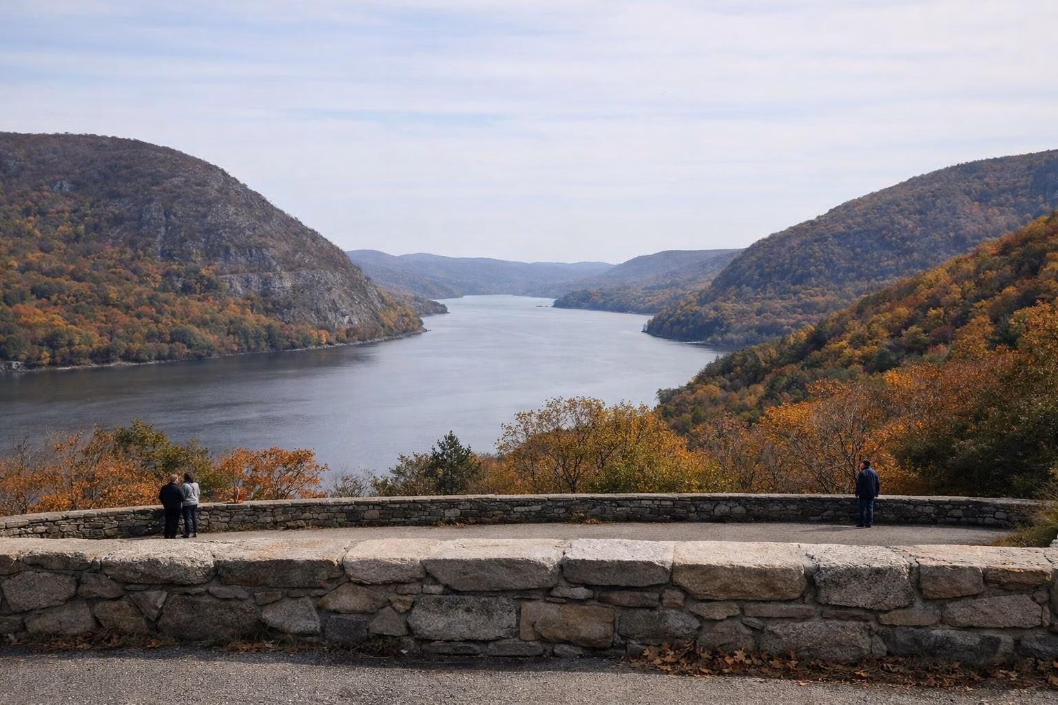 A scenic overlook with a low stone wall looking across a wide river and forested mountains in fall color, a few distant visitors